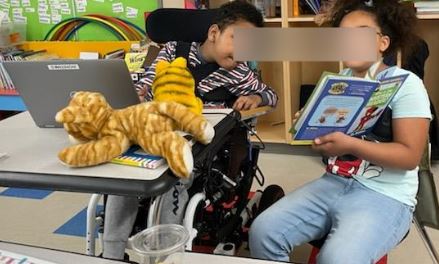A boy in a wheelchair with a girl who is holding a book.